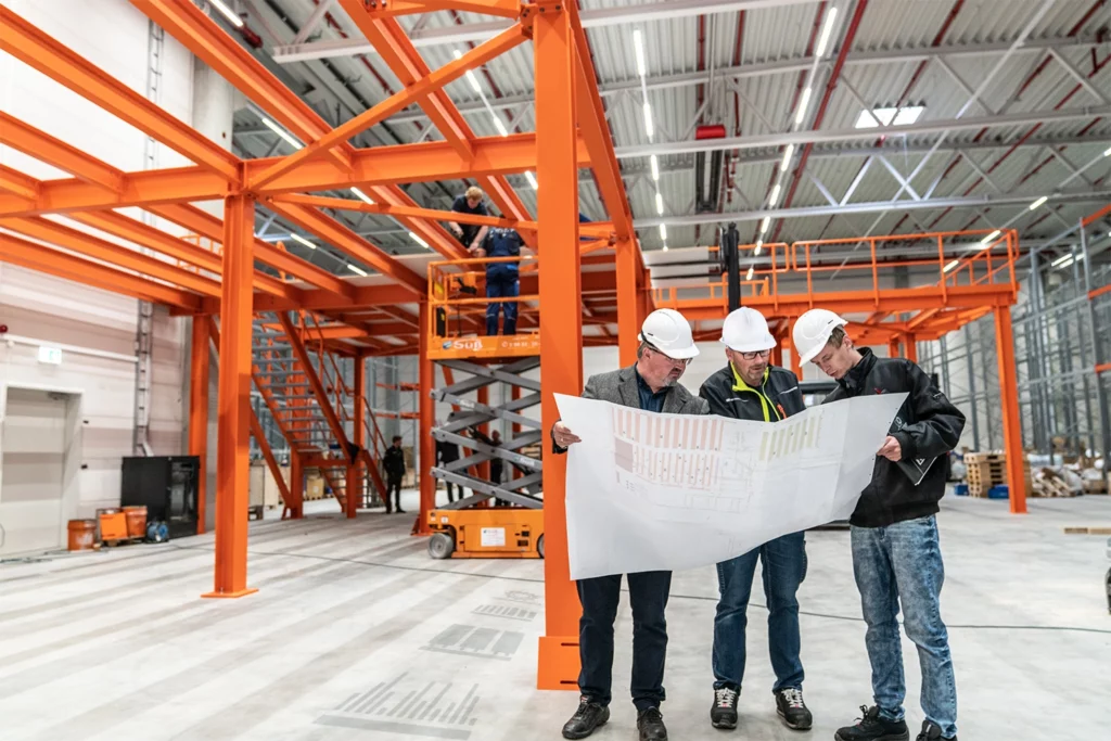 Drei Männer mit Bauplan vor einer orangefarbenen Stahlbühne